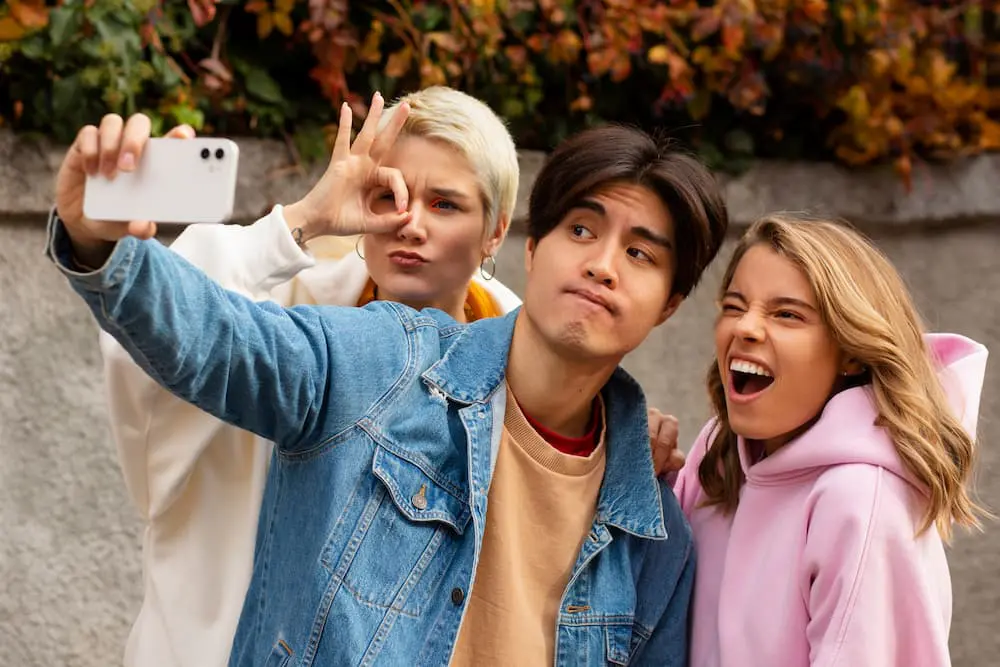 Three young adults pose for a selfie outdoorsโone makes an 'okay' gesture, another looks serious, and the third smiles with mouth openโpossibly illustrating different types of braces, with autumn leaves in the background in Jacksonville, FL.