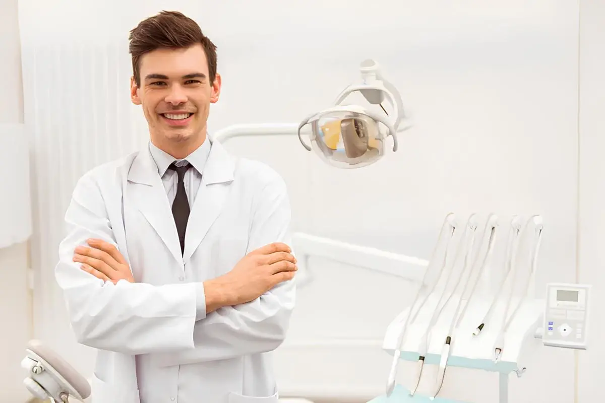 A male dentist in a white coat stands with arms crossed, smiling in a modern dental office, possibly discussing options for jaw surgery in Jacksonville, FL.