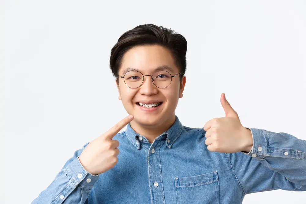 Young person wearing glasses and adult braces smiles, points to their braces with one hand, and gives a thumbs up with the other. Wears a denim shirt in Jacksonville, FL.
