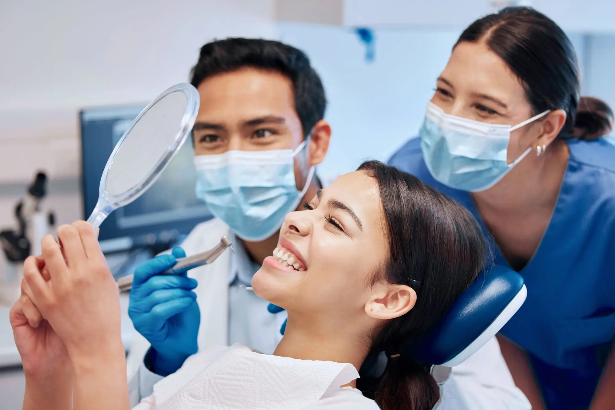 A young woman sits in a dental chair, smiling at her orthodontic appliances visible in her reflection in a handheld mirror, while two dental professionals wearing masks stand beside her in Jacksonville, FL.