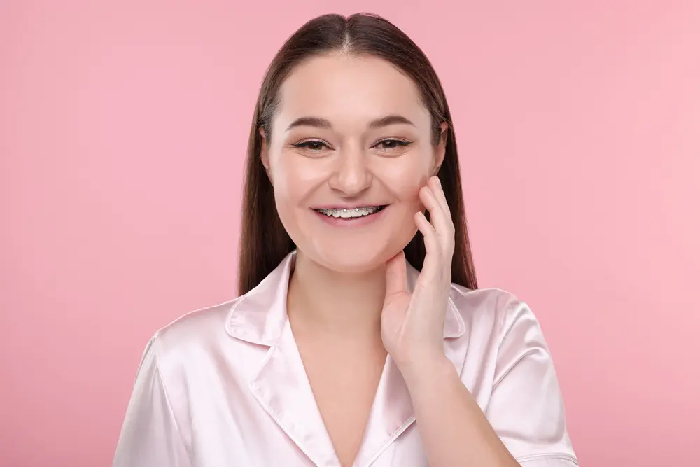 A young woman with straight brown hair wearing a light pink satin shirt smiles and touches her face, showing visible metal braces on her teeth, in front of a pink background in Jacksonville, FL.