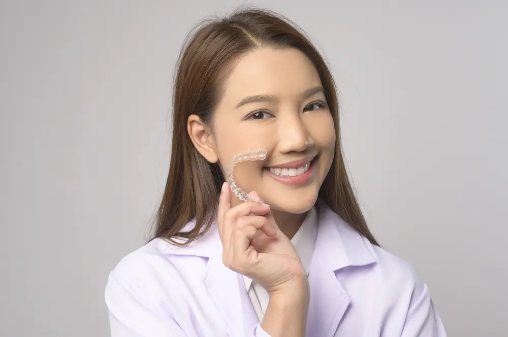 Orthodontic Insurance Plans: A woman in a lab coat smiles and holds a clear dental aligner in front of her face against a plain background in Jacksonville, FL.