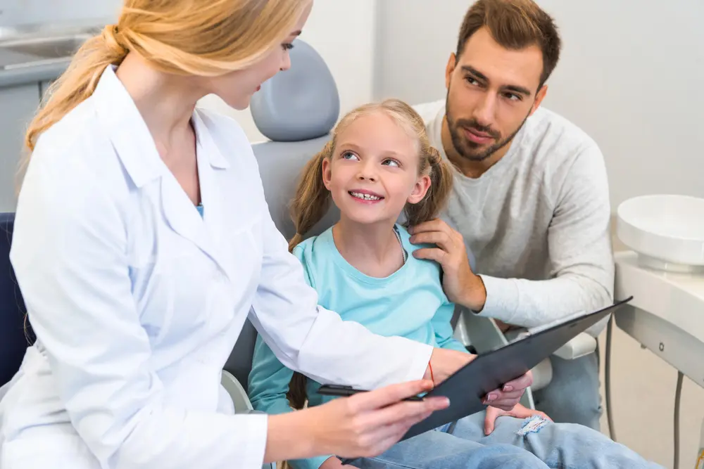 A female dentist talks to a young girl about children orthodontics in a dental chair while her father sits beside her, gently holding her shoulder in Jacksonville, FL.
