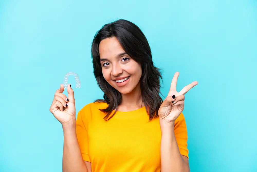 Orthodontic Insurance Plans: Woman in a yellow shirt smiles, holding a clear dental aligner in one hand and making a peace sign with the other, in front of a light blue background in Jacksonville, FL.