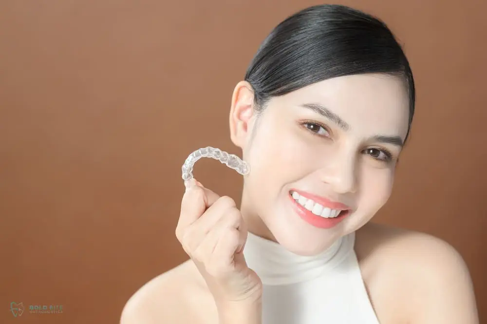 A woman smiles while holding a clear dental aligner near her face against a plain brown background after understanding "Does Insurance Cover Invisalign in Jacksonville, FL".