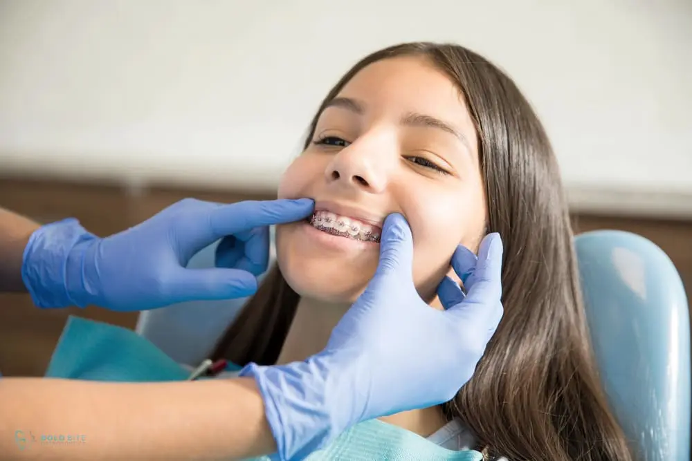 During a dental check-up at Bold Bite Orthodontics in Jacksonville, FL, a dentist wearing blue gloves examines a young girl's braces showcasing the Invisalign vs Braces cost in Jacksonville, FL at Bold Bite Orthodontics.