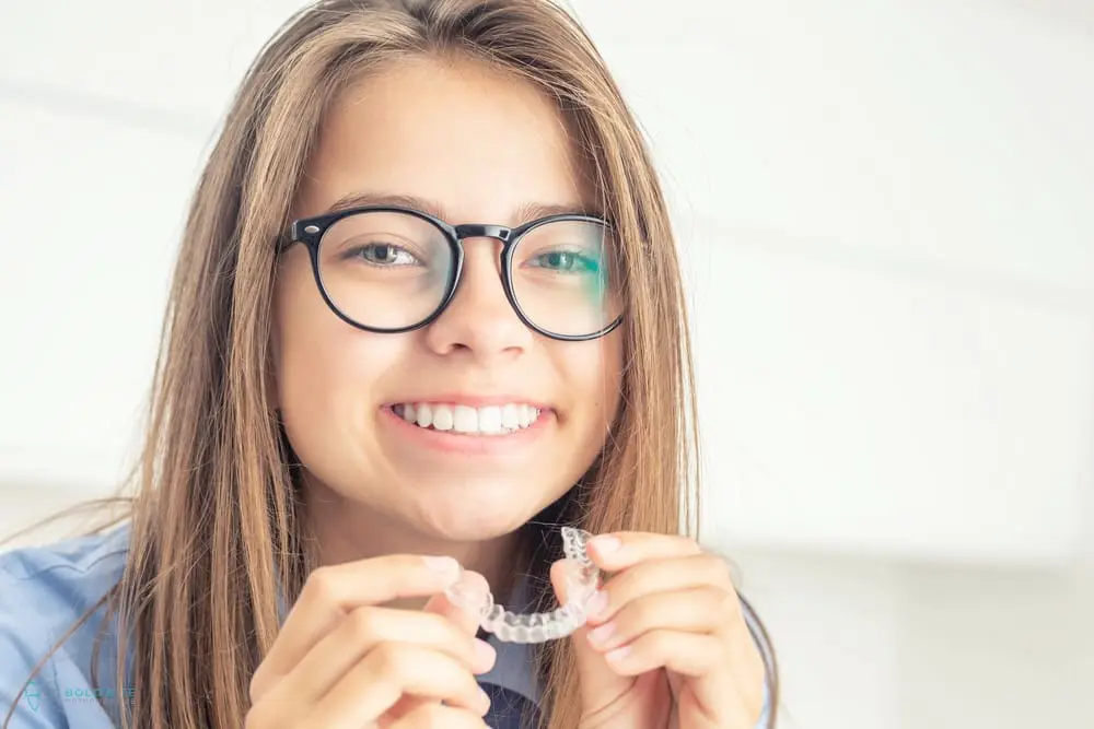 A smiling young person with glasses holds a clear dental aligner in front of their mouth, showcasing the Invisalign vs Braces cost in Jacksonville, FL at Bold Bite Orthodontics. 