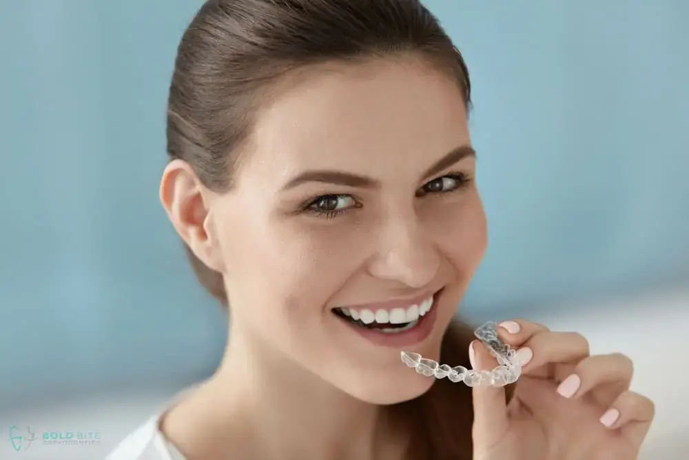 A woman smiles, holding a clear aligner near her mouth at Bold Bite Orthodontics in Jacksonville, FL, curious about Invisalign results - How Long Does Invisalign Take in Jacksonville, FL. A woman smiles, holding a clear aligner near her mouth at Bold Bite Orthodontics in Jacksonville, FL, curious about Invisalign results - How Long Does Invisalign Take in Jacksonville, FL.