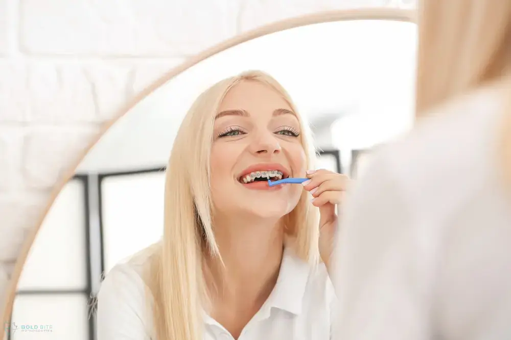 Brushing her teeth in front of a round mirror, a woman with long blonde hair at Bold Bite Orthodontics in Jacksonville, FL - How to Floss with Braces in Jacksonville, FL Brushing her teeth in front of a round mirror, a woman with long blonde hair at Bold Bite Orthodontics in Jacksonville, FL - How to Floss with Braces in Jacksonville, FL