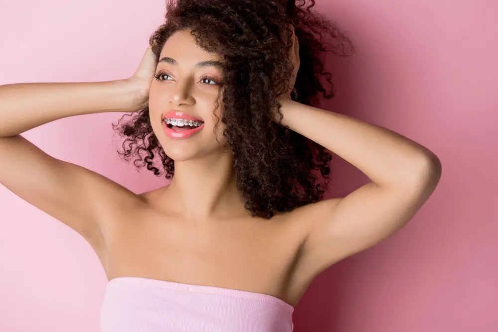 Smiling woman with curly hair and braces poses in a pink top, representing Bold Bite Orthodontics in Jacksonville, FL - How To Get Braces Glue Off Teeth in Jacksonville, FL