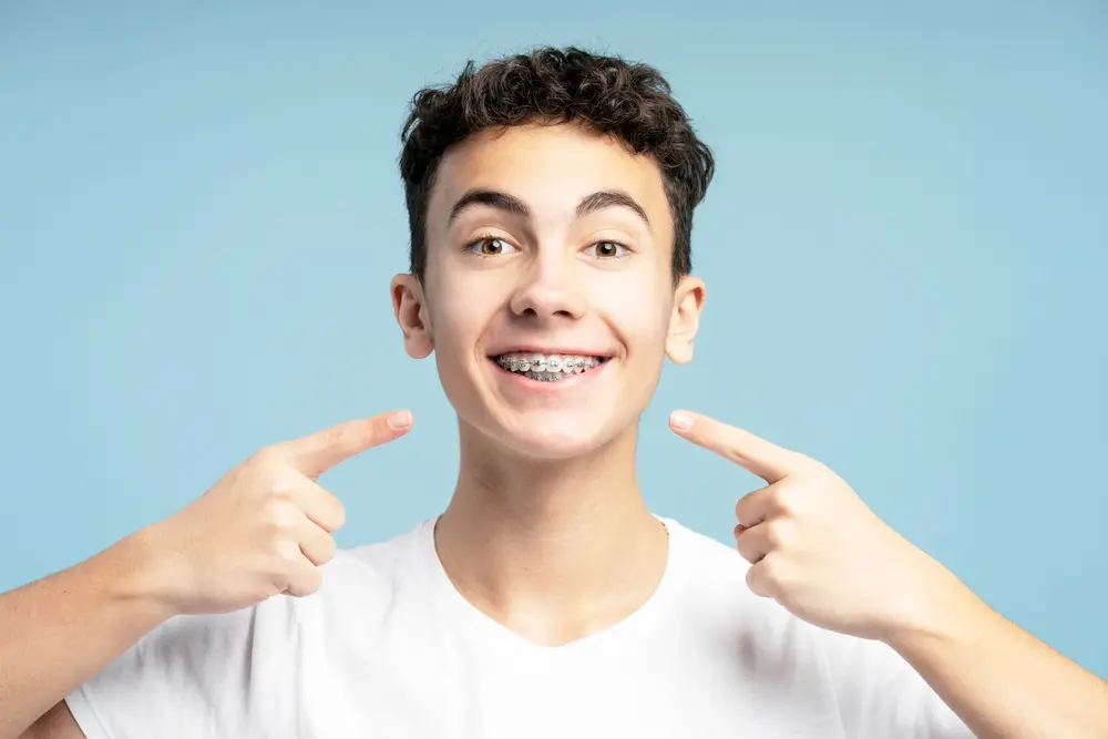 A teen in a white t-shirt smiles, pointing to braces at Bold Bite Orthodontics in Jacksonville, FL, with a blue background - How to Clean Braces in Jacksonville, FL