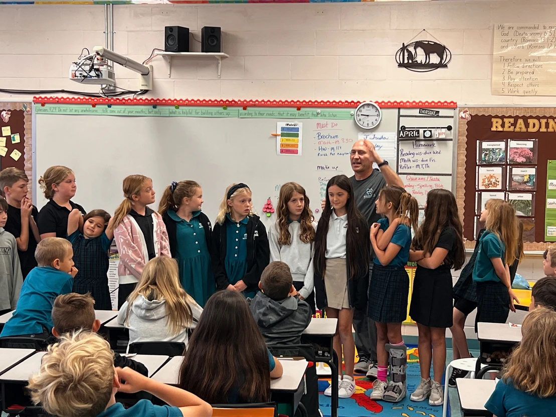 Dr. Marty Greenberg, orthodontist expert at Bold Bite Orthodontics in Jacksonville FL meet with a group of students stands at the front of a classroom, lined up in a row. An adult stands with them, speaking. Several students sit at desks, facing the group.