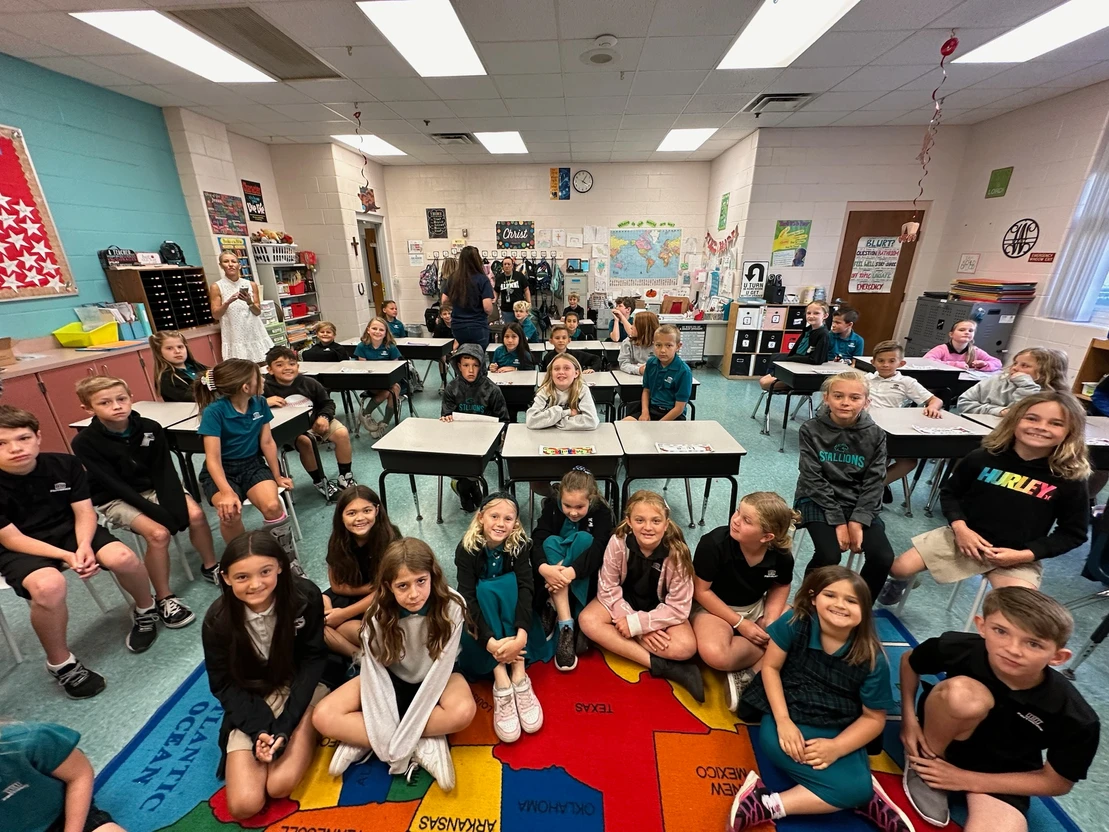 Dr. Marty Greenberg, orthodontist expert at Bold Bite Orthodontics in Jacksonville FL meet with a classroom with 32 children sitting on chairs and a colored map carpet, facing the camera. The room has educational posters, maps, and school supplies. Two adults stand in the back.