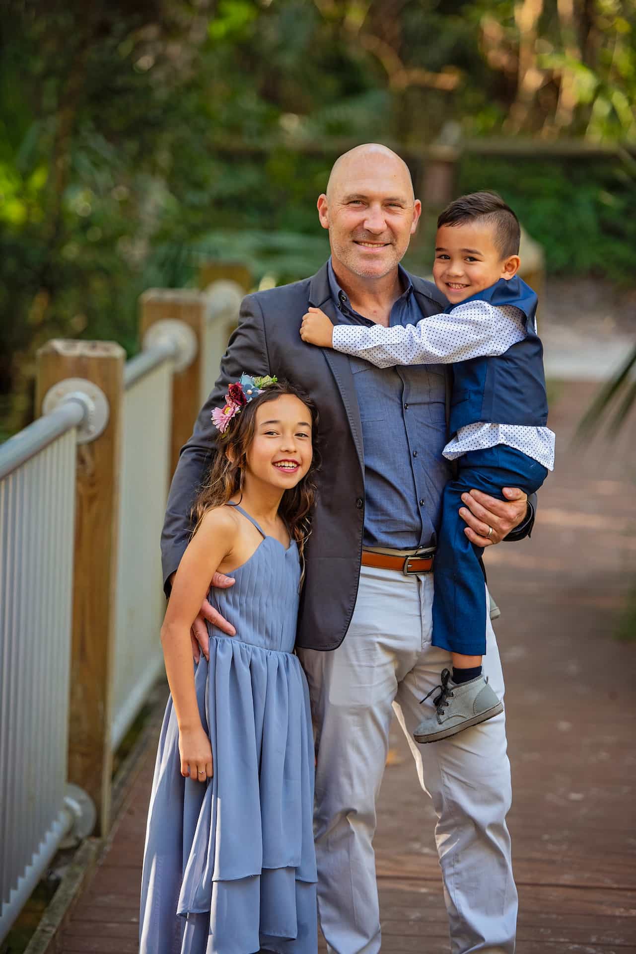 Dr. Marty Greenberg stands on a wooden path outdoors, smiling with a girl in a blue dress and a boy in a vest and tie, both joyfully embracing him in Jacksonville, FL