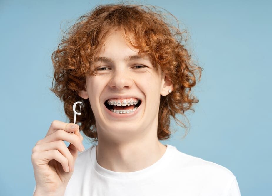 A smiling red-haired teen with braces holds dental floss pick at Bold Bite Orthodontics in Jacksonville, FL, blue background - How to Floss with Braces in Jacksonville, FL